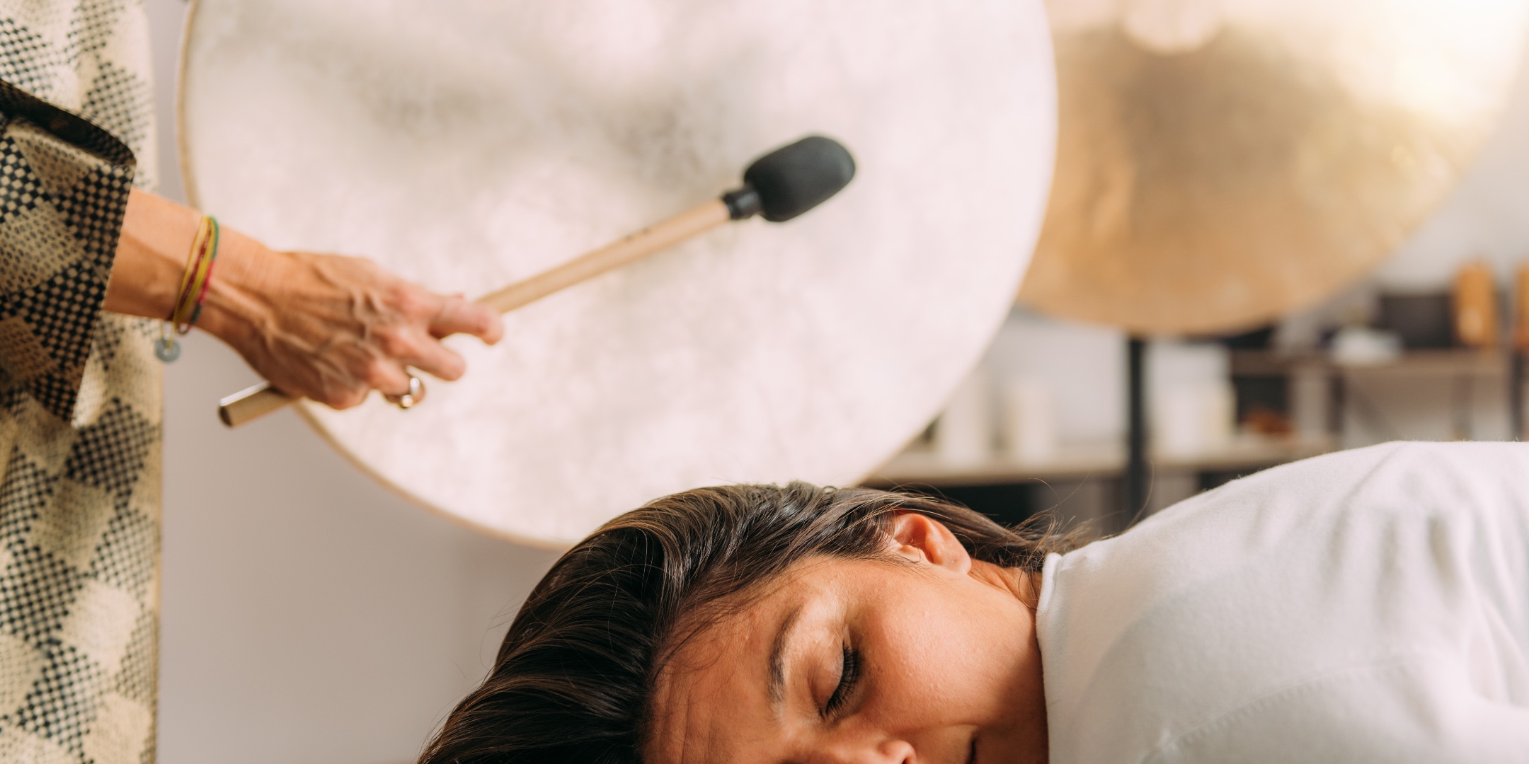 Sound healing practitioner playing a frame drum beside a symphonic gong during a gong bath session, with a client resting nearby